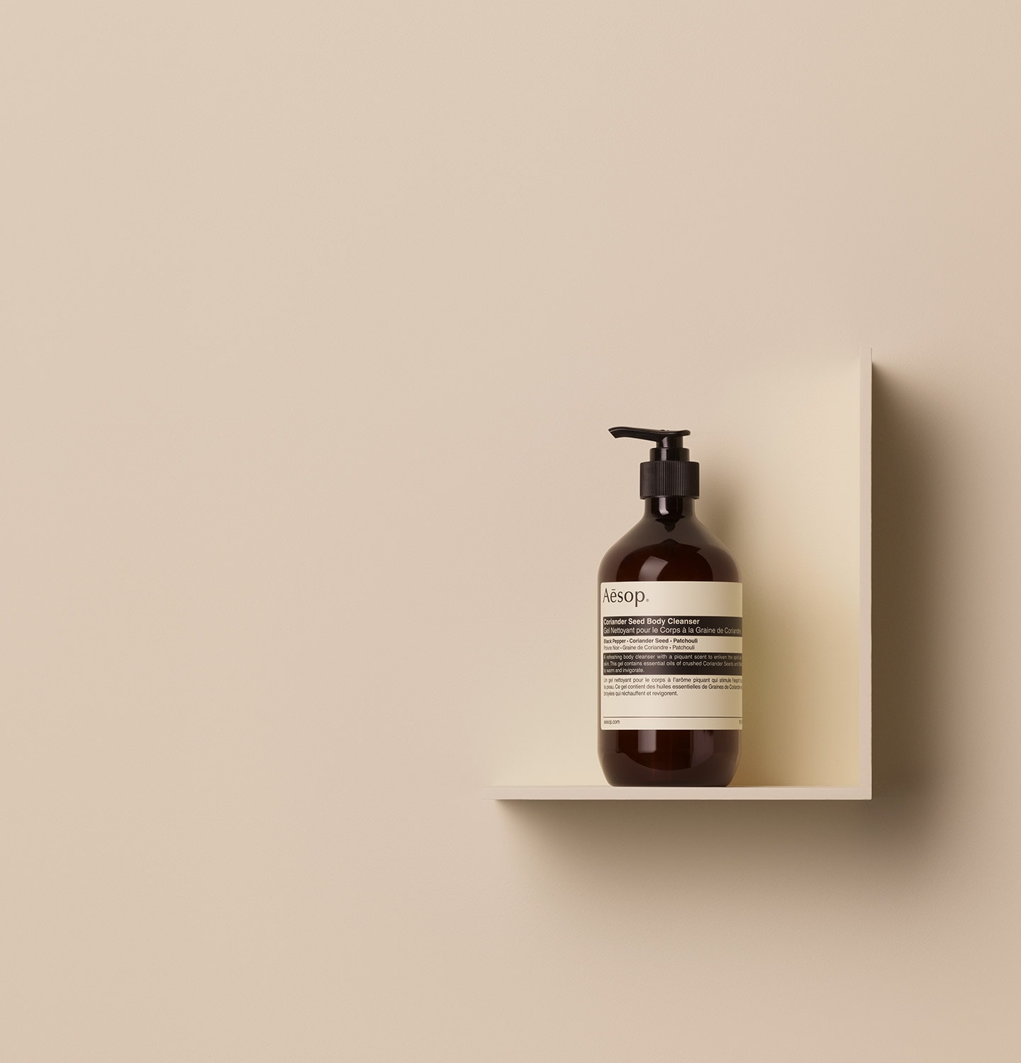 Aesop Coriander Seed Body Cleanser in 500ml amber pump bottle placed on a shelf in a beige surface