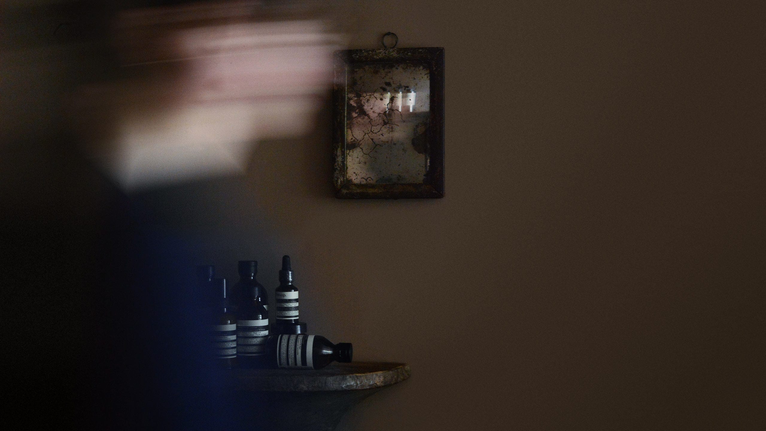 Blurred figure moving in front of an age-spackled mirror and collected Aesop products in background.