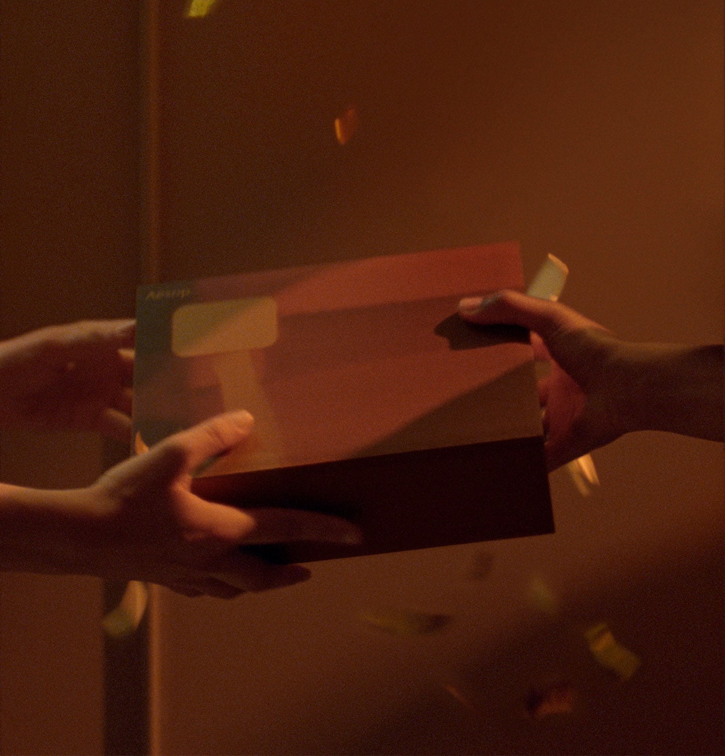 Brown-orange hued photo of hands exchanging a gift box. Confetti is falling from above.